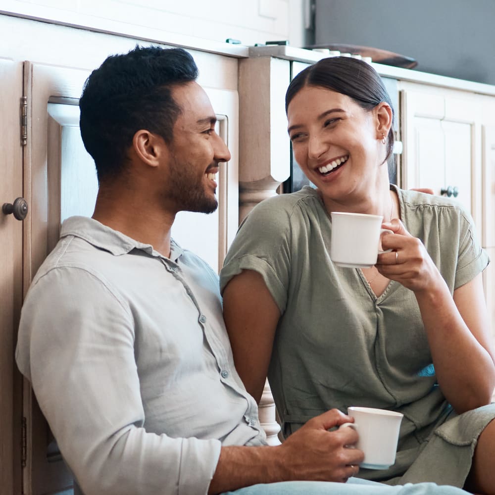 A couple is sitting on the floor of a kitchen smiling at each other. They are both holding mugs.